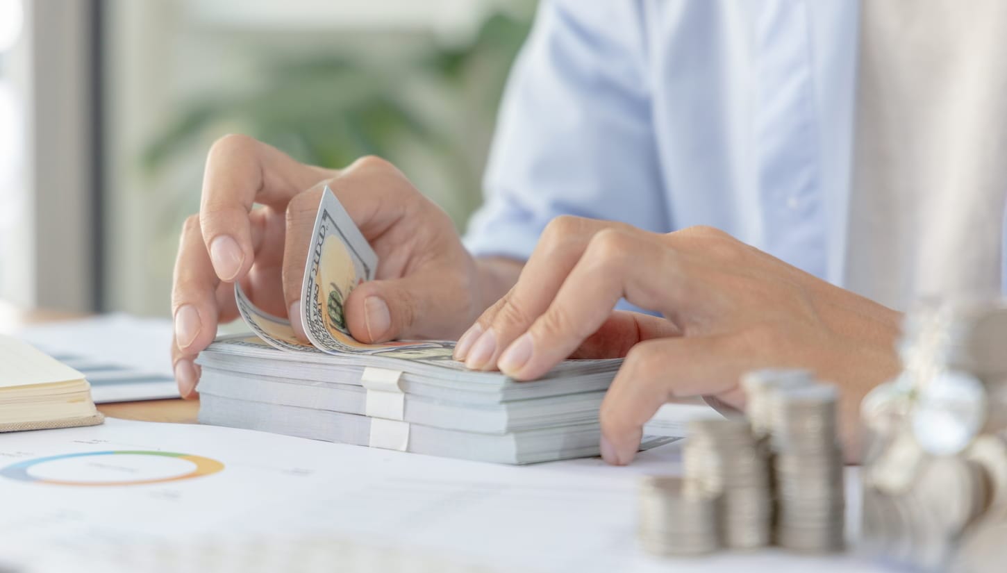 An image of a Businessman's hands counting 100 dollar bills, and coins are stacked on top of the desk.