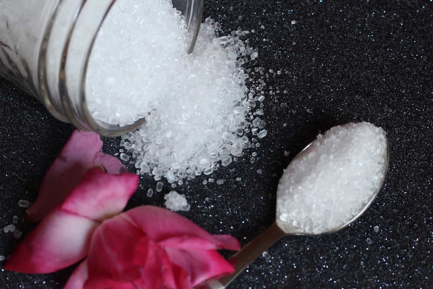 An image of Epsom salt in an opened bottle scattered on a black tiled countertop. A spoon full of Epsom salt and rose petals on the side.
