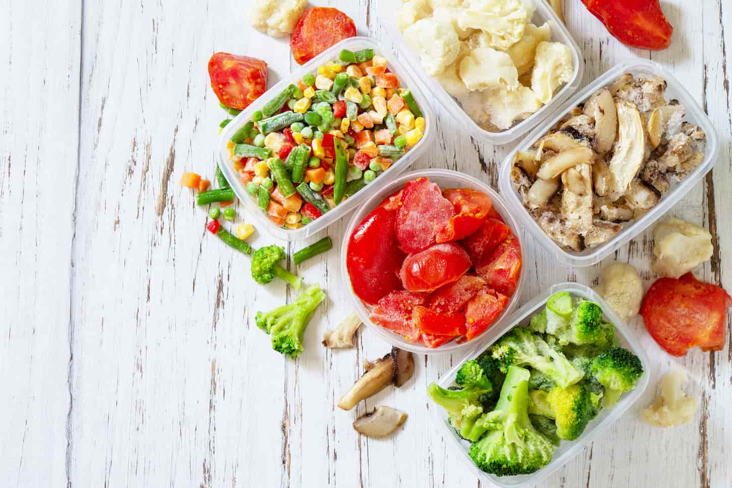 An image of multiple containers with frozen vegetables and mushrooms on a white wooden table.