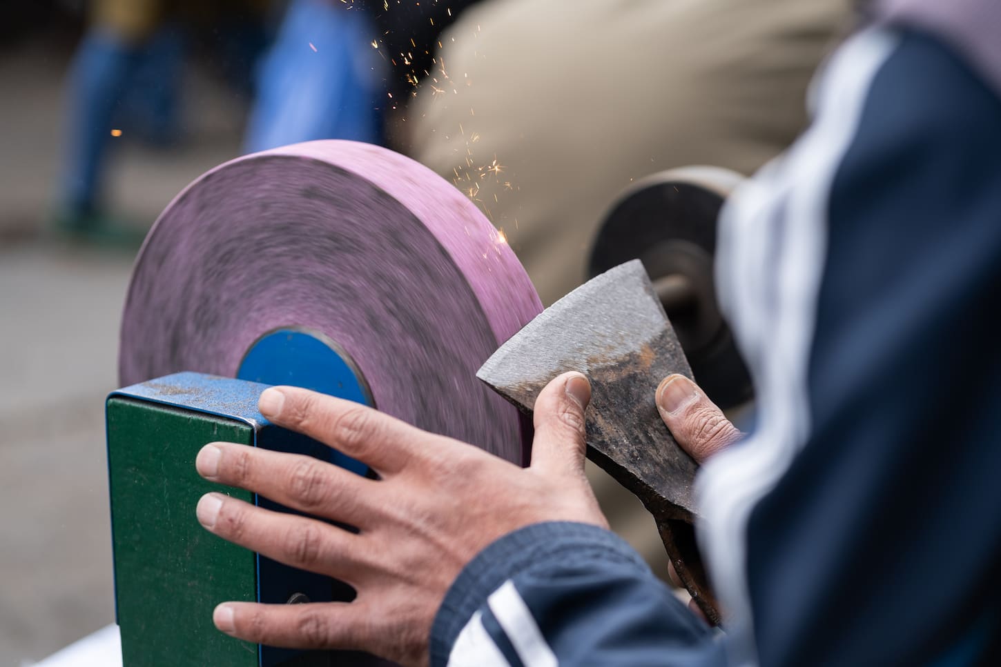 An image of a Master sharpening axe on an electric sharpener with bright sparks and holding a blade with his brutish fingers.