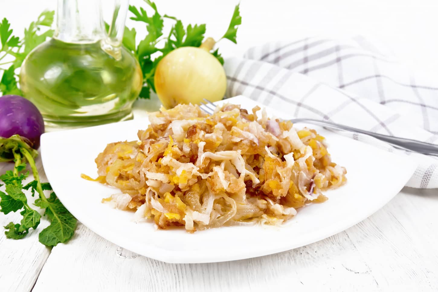 An image of fried turnips with onions on a plate, oil in a decanter, and napkin on the background of a light wooden board.