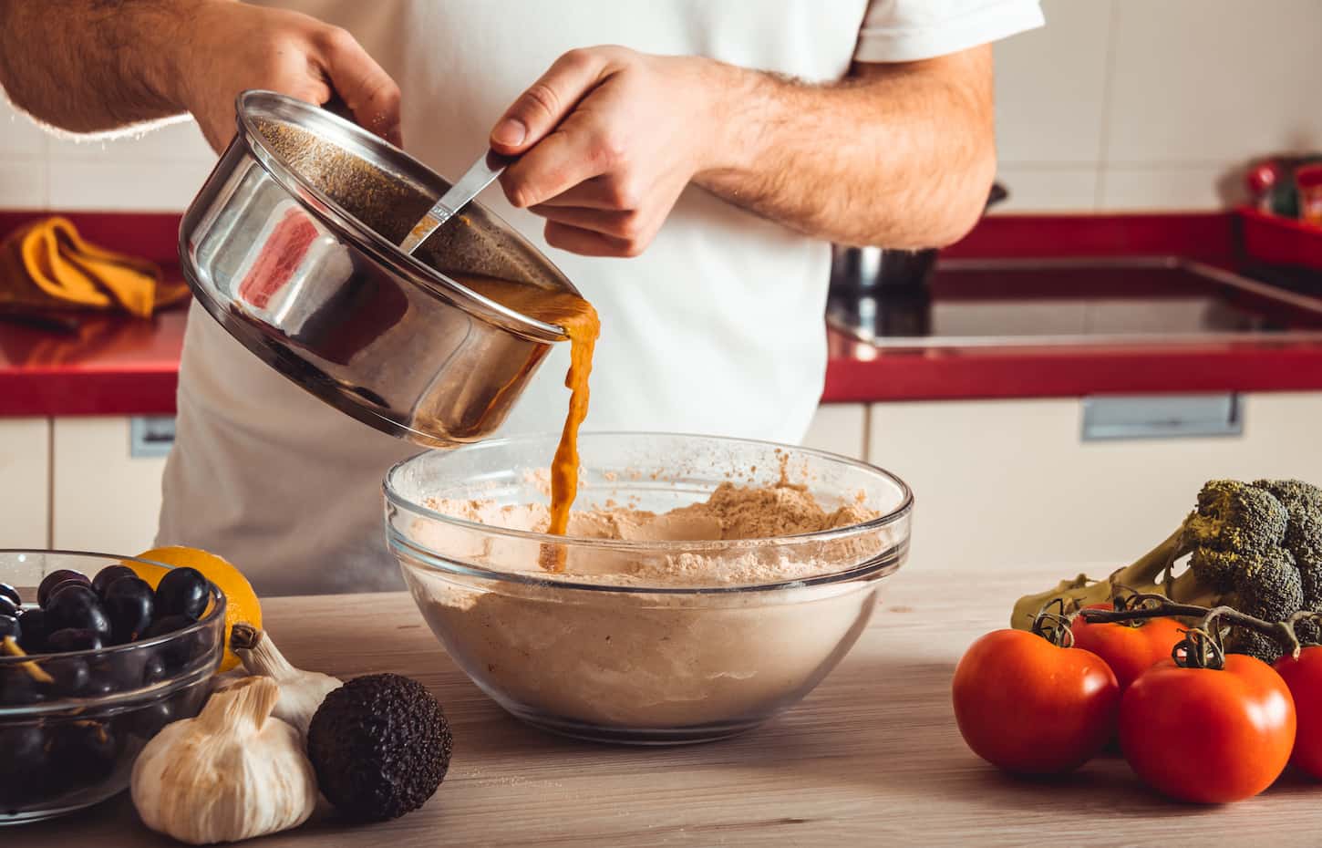An image of a man's hands while mixing wheat flour on a clear bowl using a spoon, with tomatoes, garlic,, and broccoli on top of a light brown wooden table.