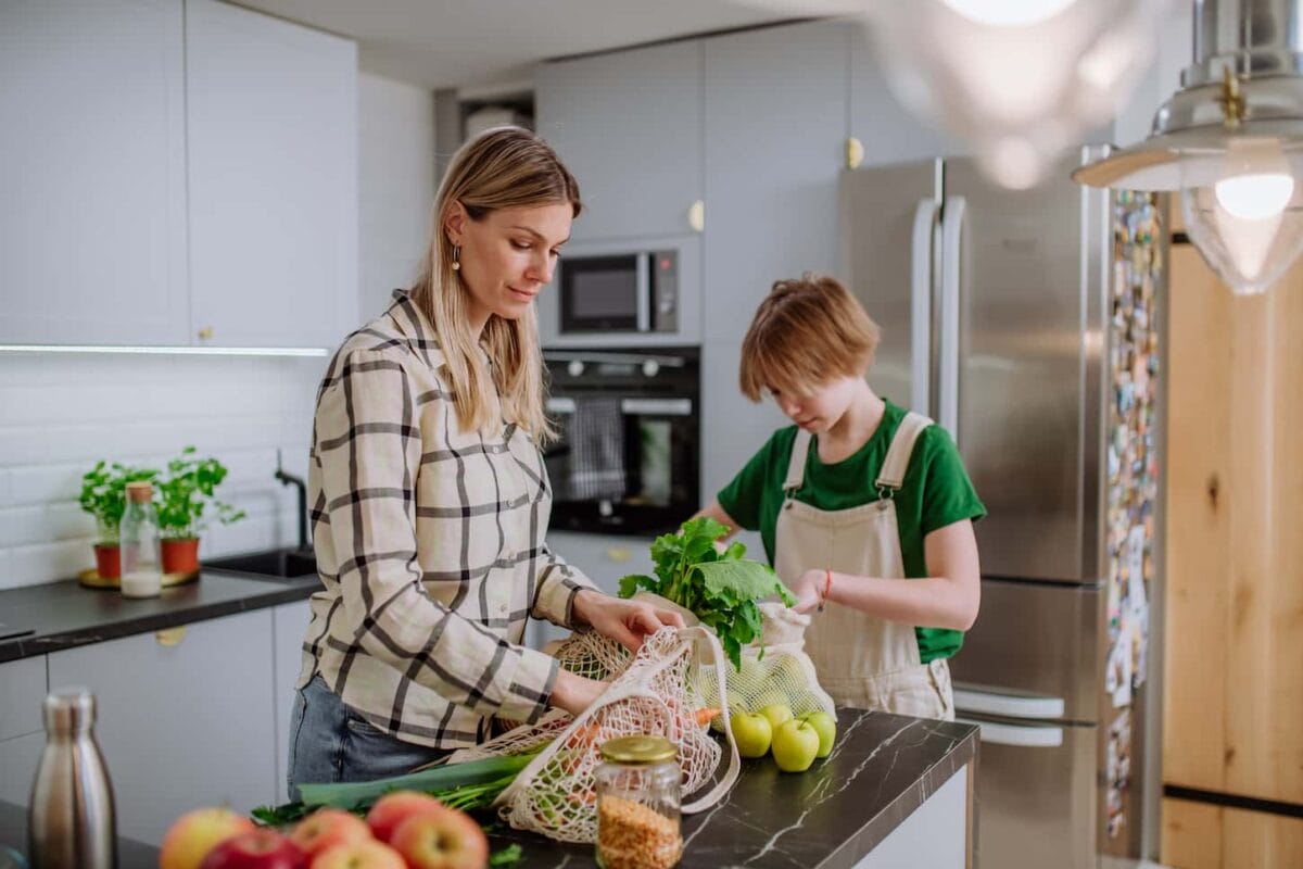 An image of A mother unpacking local food in zero-waste packaging from a bag with help of her daughter in the kitchen at home.