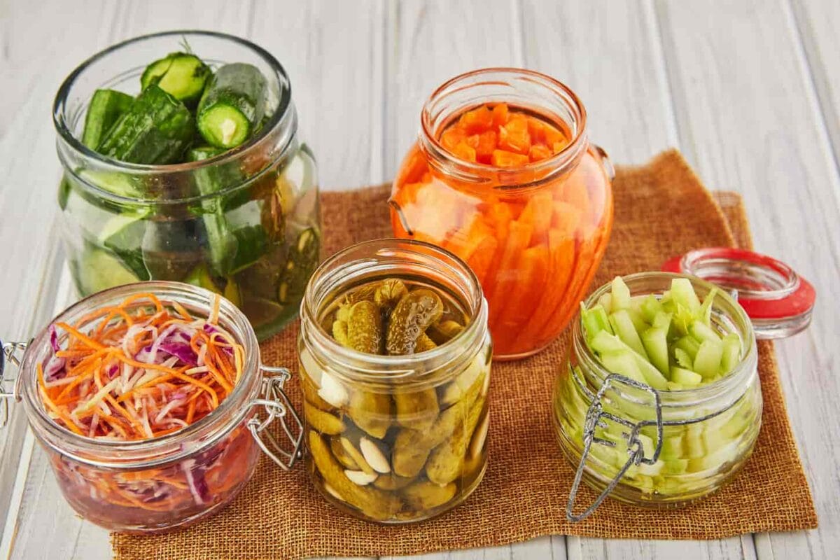 An image of fermented Sour sauerkraut, pickled carrots, pickled cucumbers, and pickled celery in glass jars on a white wooden kitchen table.